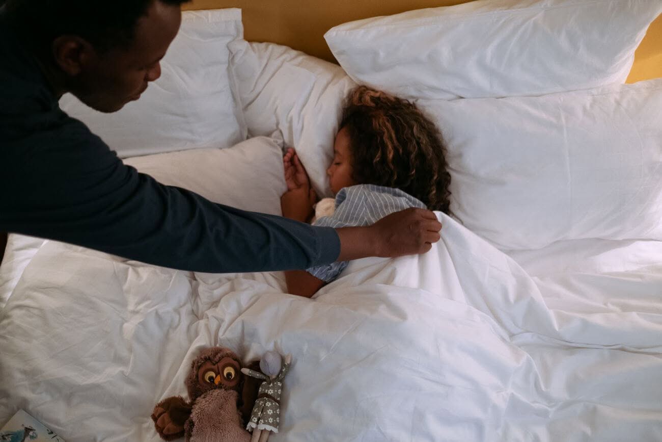 Una babysitter rimbocca le coperte a una bambina che dorme, con luce soffusa